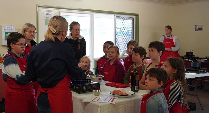 A teacher at the Charleville ScA group of young students wearing red aprons listening to a chef talking about food and food preparation as part of Jamie Ministry of Food cooking program during school holidays.